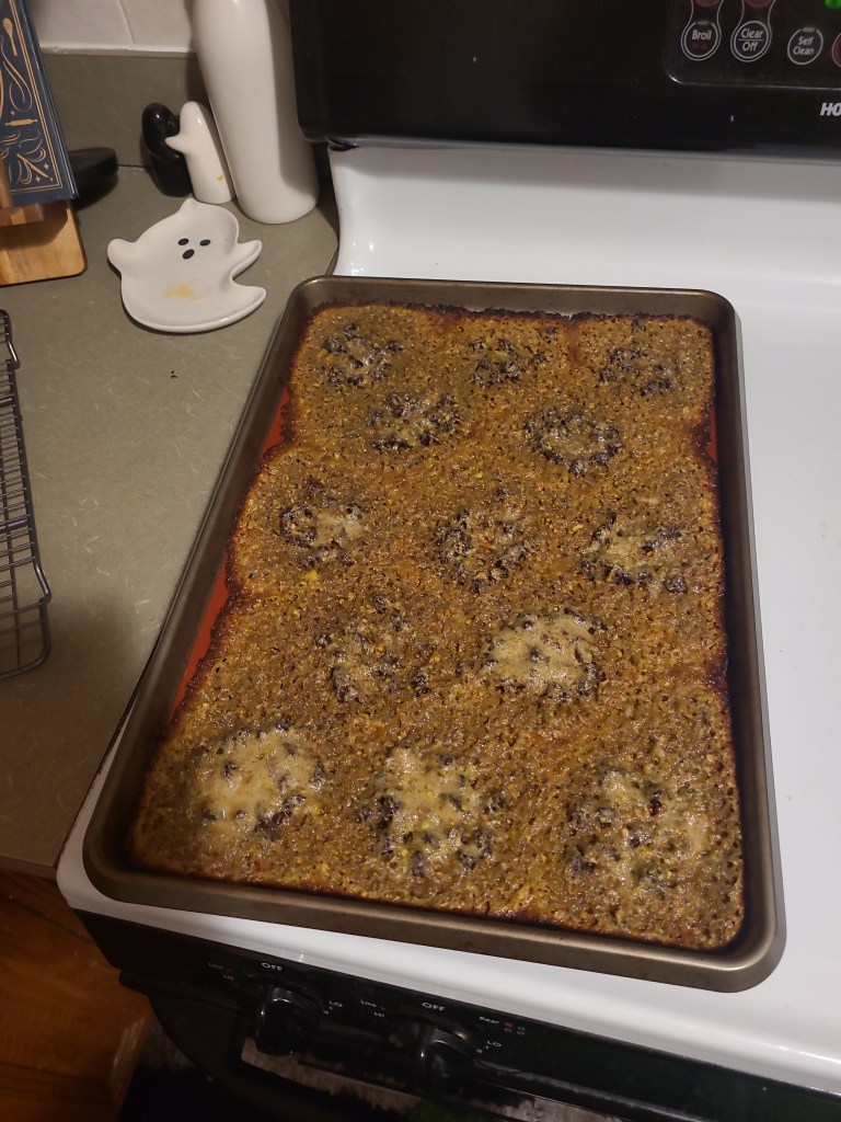 a cookie sheet sitting on top of a stove. On the cookie sheet are 13 cookies that are identifiable via distinct lumps at various points but the edges have melted and blended together to cover the entire sheet. 