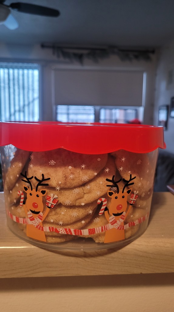 Many snickerdoodle cookies in a clear plastic container decorated with cute reindeer holding presents and candy canes. The container has a red lid.